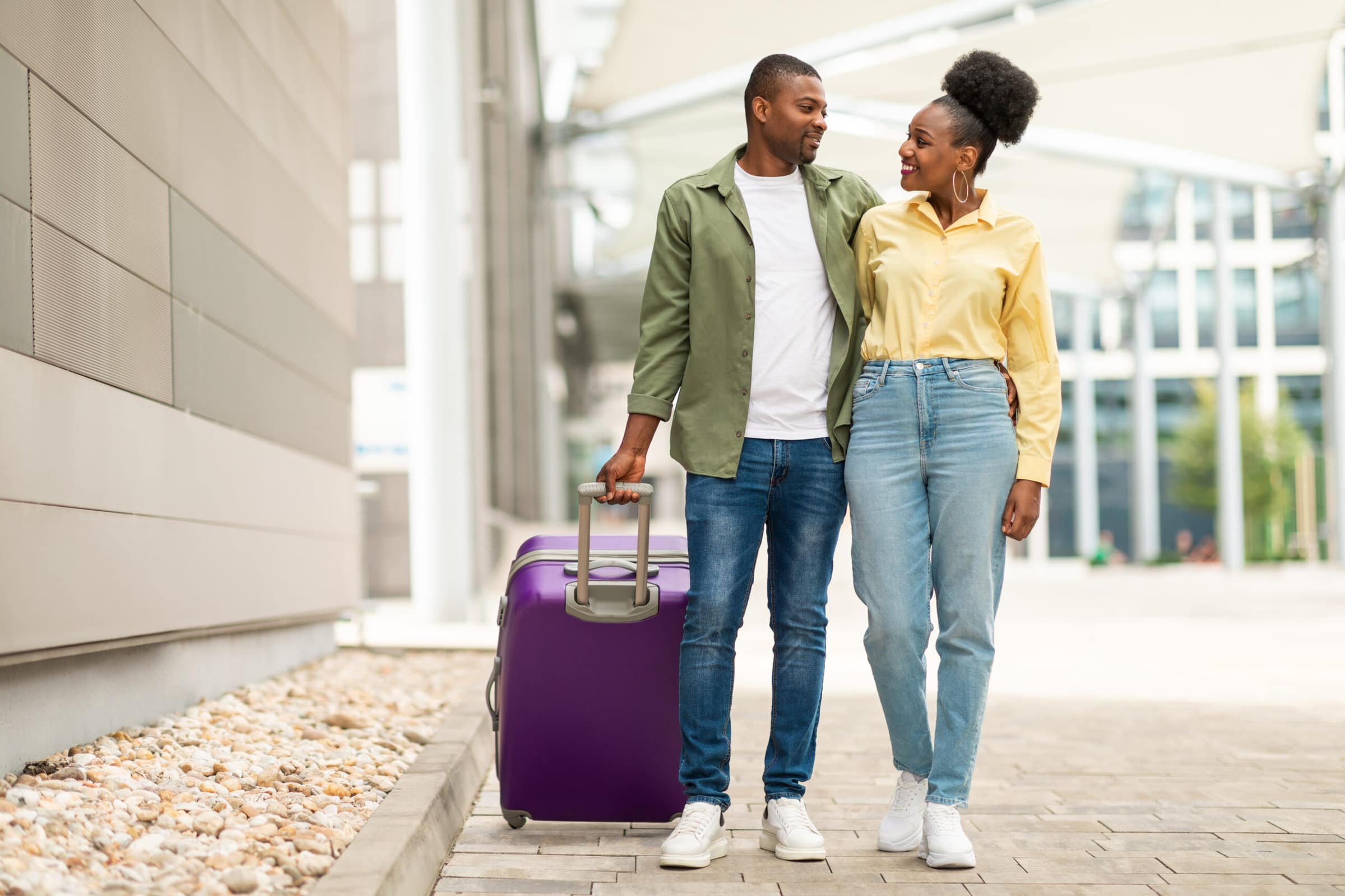 Cheerful African American Spouses With Travel Suitcase Standing Smiling To Each Other Near Airport Outdoors. Full Length Shot Of Tourists Couple Having Vacation Trip. Tourism And Transportation
