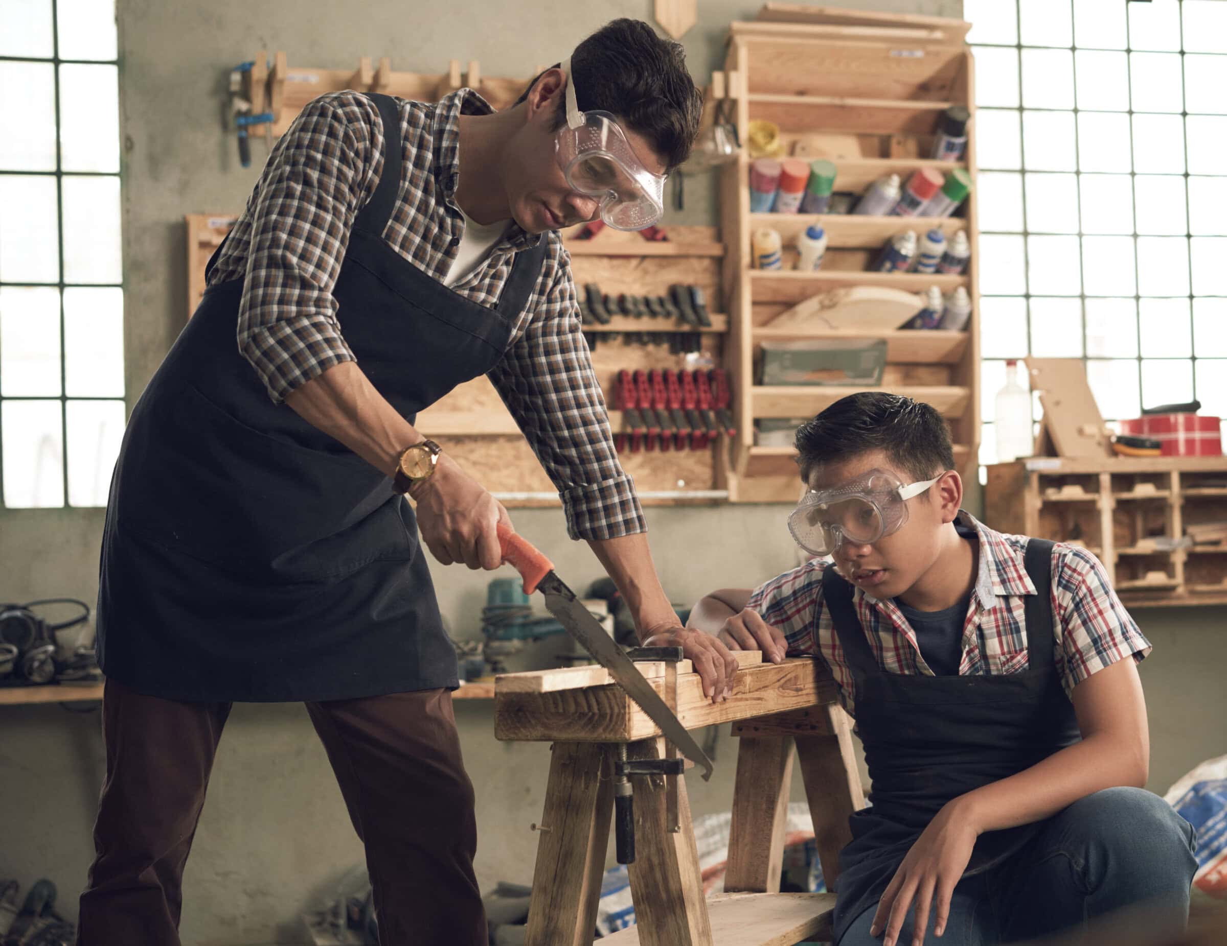 Vietnamese teenage boy looking how his father sawing wooden plank