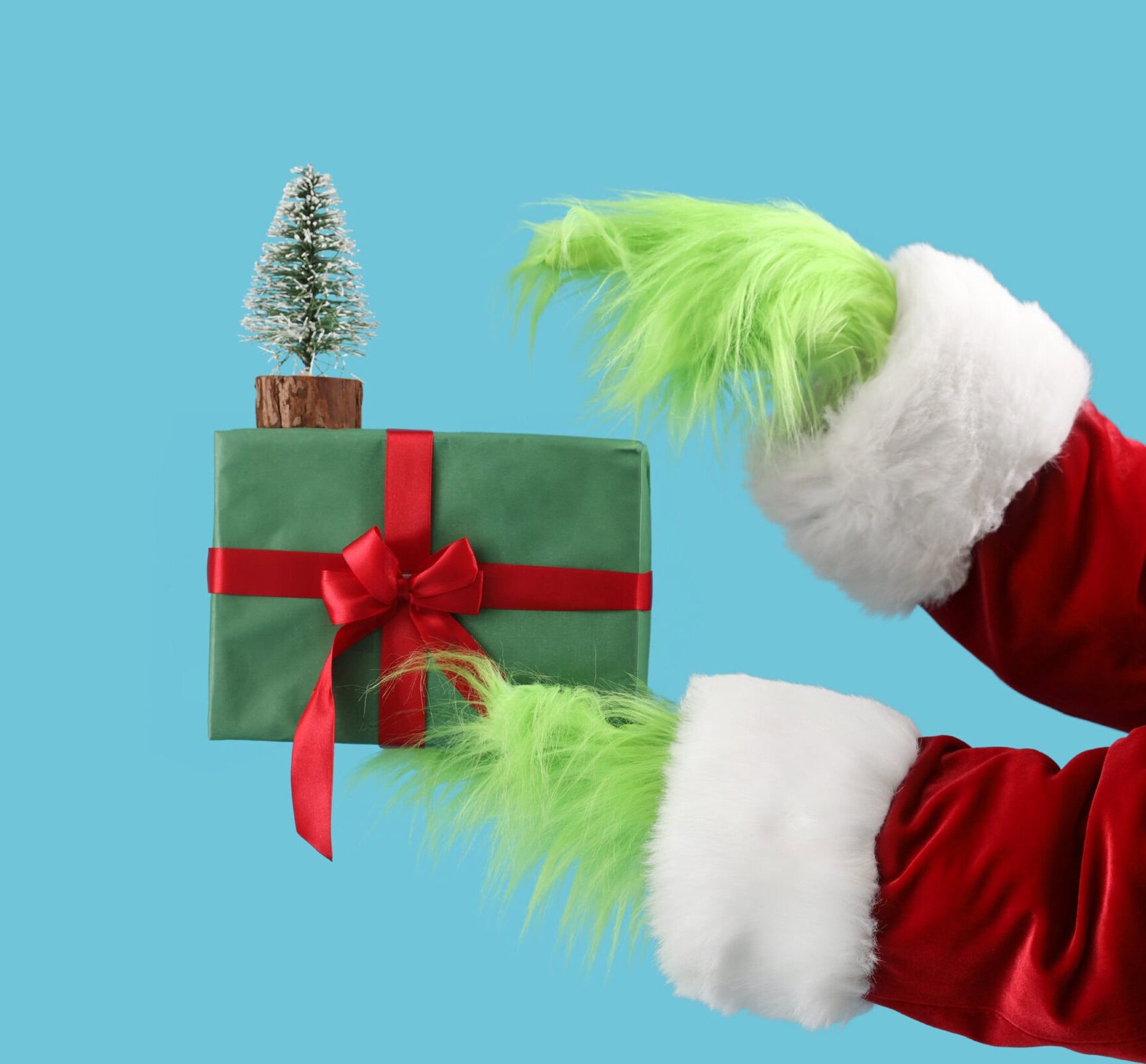 Green hairy hands of creature in Santa costume with decorative fir tree and Christmas gift box on blue background