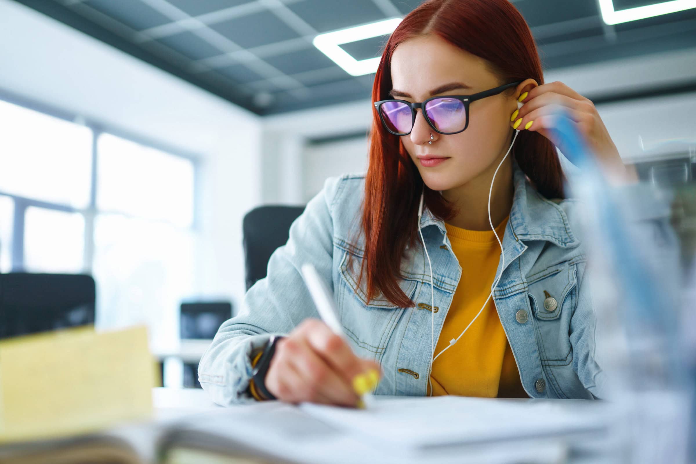 Portrait of girl works at a computer in the modern office. Young freelancer working from a laptop computer. Teenager student at the computer listens music to earphones. Business woman with glasses.