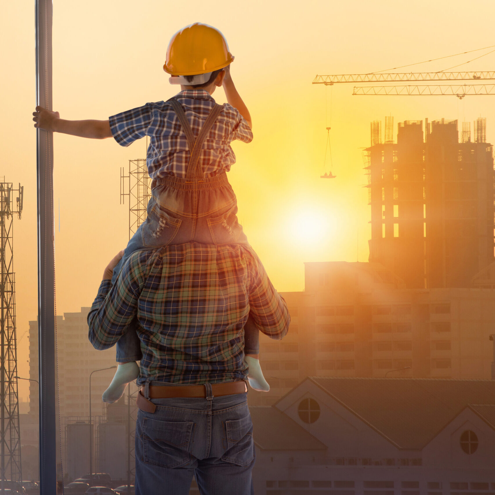 Asian boy on father's shoulders with background of new high buildings and silhouette construction cranes of evening sunset, father and son concept